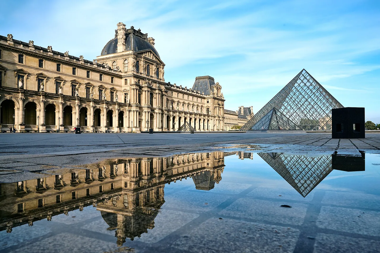 The Louvre’s palace wing and glass pyramid in an empty Paris courtyard, reflected in a rain puddle in the early morning light