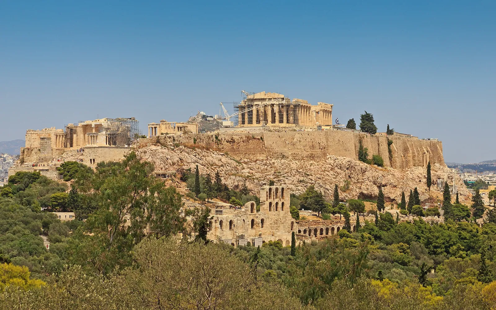 The Parthenon crowning the Acropolis hill above Athens, seen from a distance across trees and rooftops