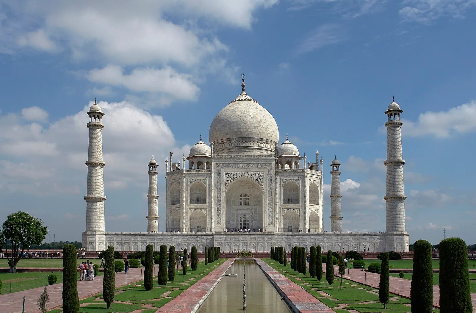 The white dome and four minarets of the Taj Mahal seen along a long central reflecting pool, with garden paths and cypress trees, under a blue sky with clouds