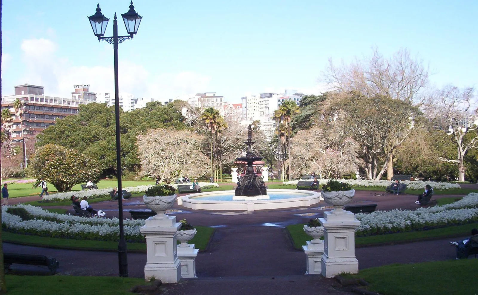 A fountain surrounded by flower beds and trees in Albert Park, with city buildings behind