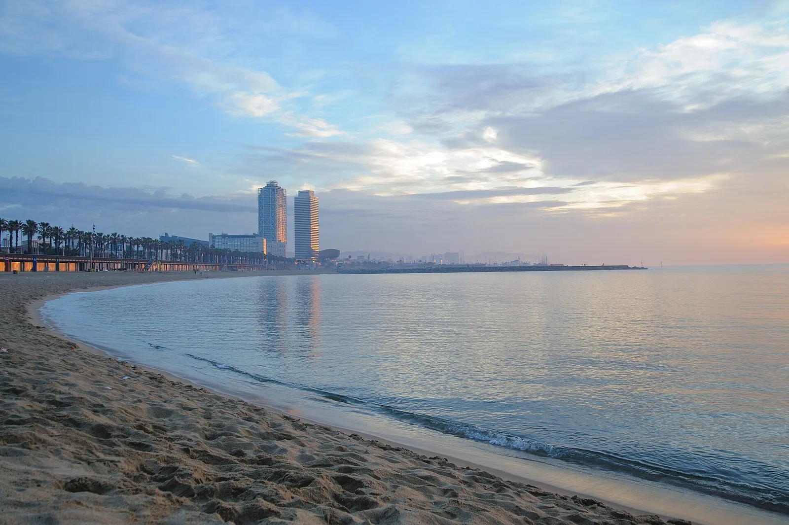 Barceloneta Beach at dawn, with calm water, pale sand, and the two towers of the Olympic Port on the horizon