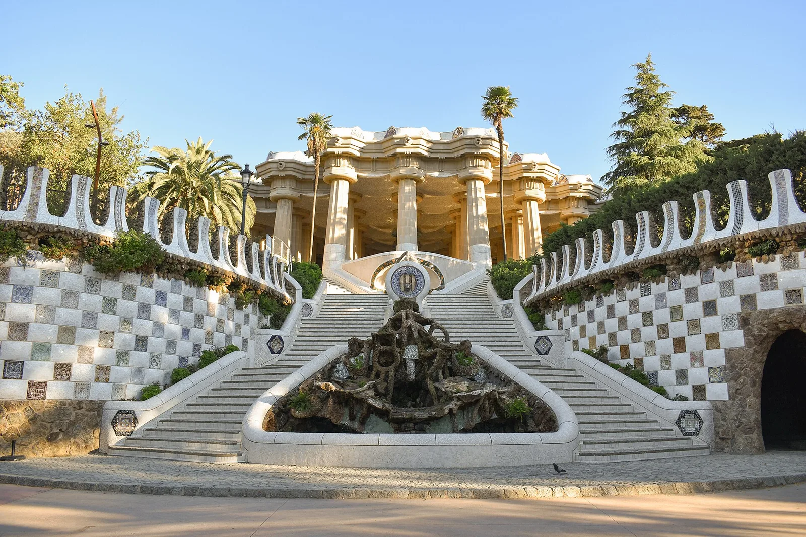 The mosaic-lined stairs at the entrance of Park Güell leading up to the columned hall, with a fountain halfway up
