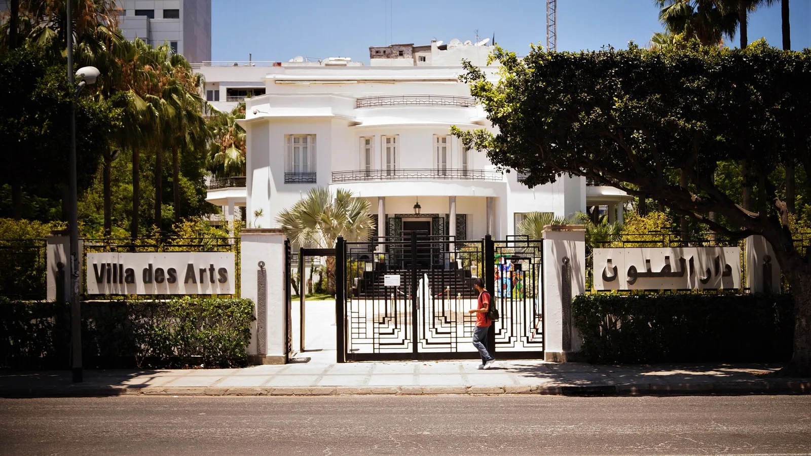 The white, Art Deco facade of the Villa des Arts behind its iron gate and palm trees