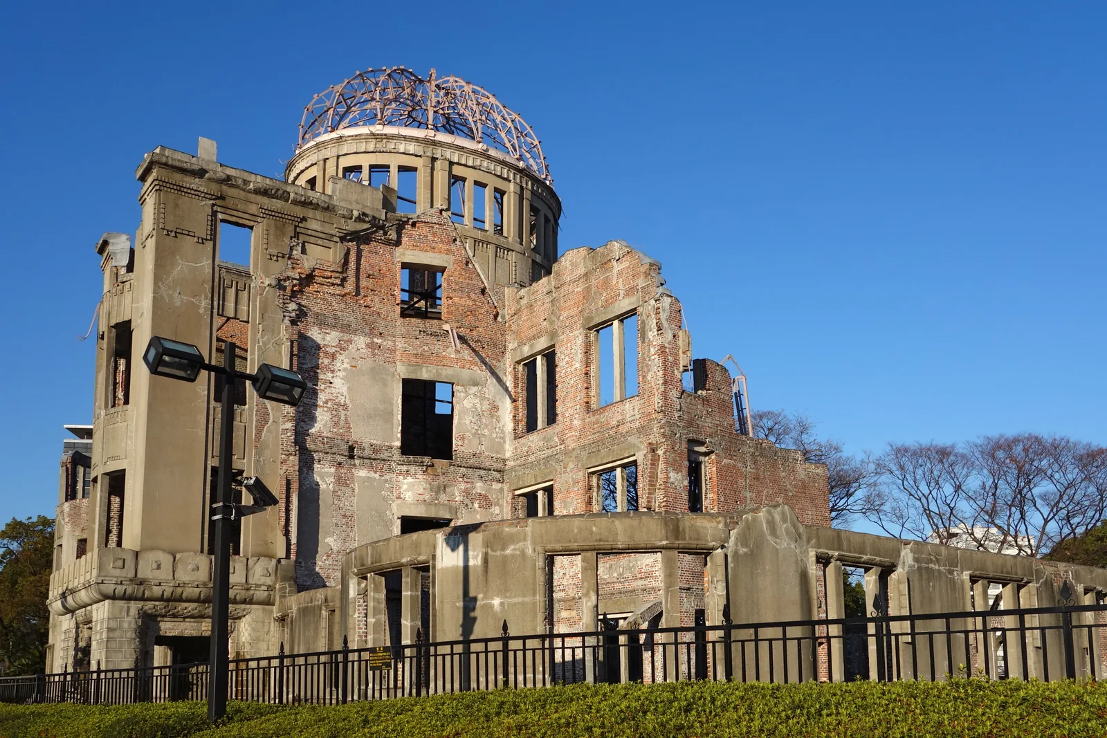 The ruined brick-and-steel dome of the Atomic Bomb Dome against a clear blue sky, seen behind an iron fence