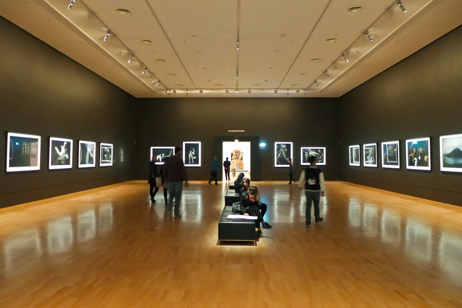 A gallery hall at the National Gallery of Victoria, with lit photographs on dark walls and visitors on a central bench