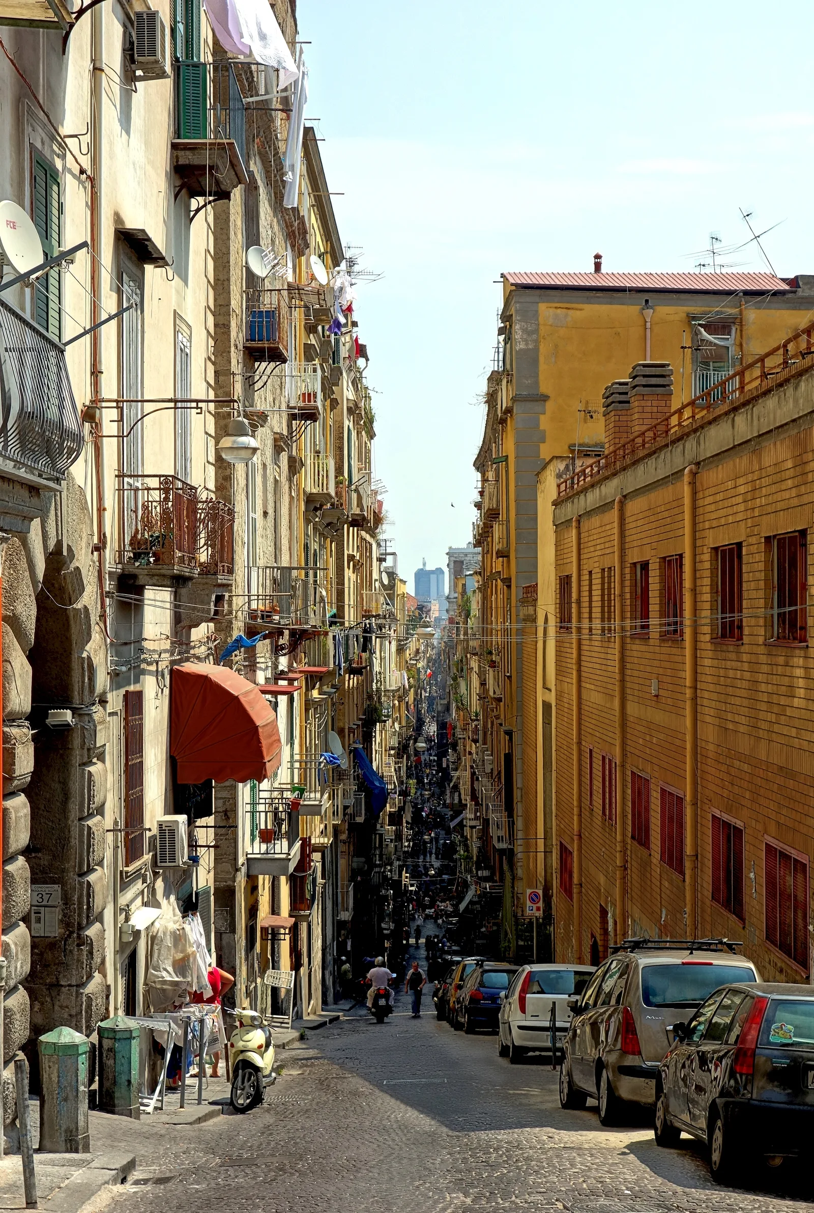 A narrow, steep Naples street lined with balconies, laundry, and scooters, looking toward the city