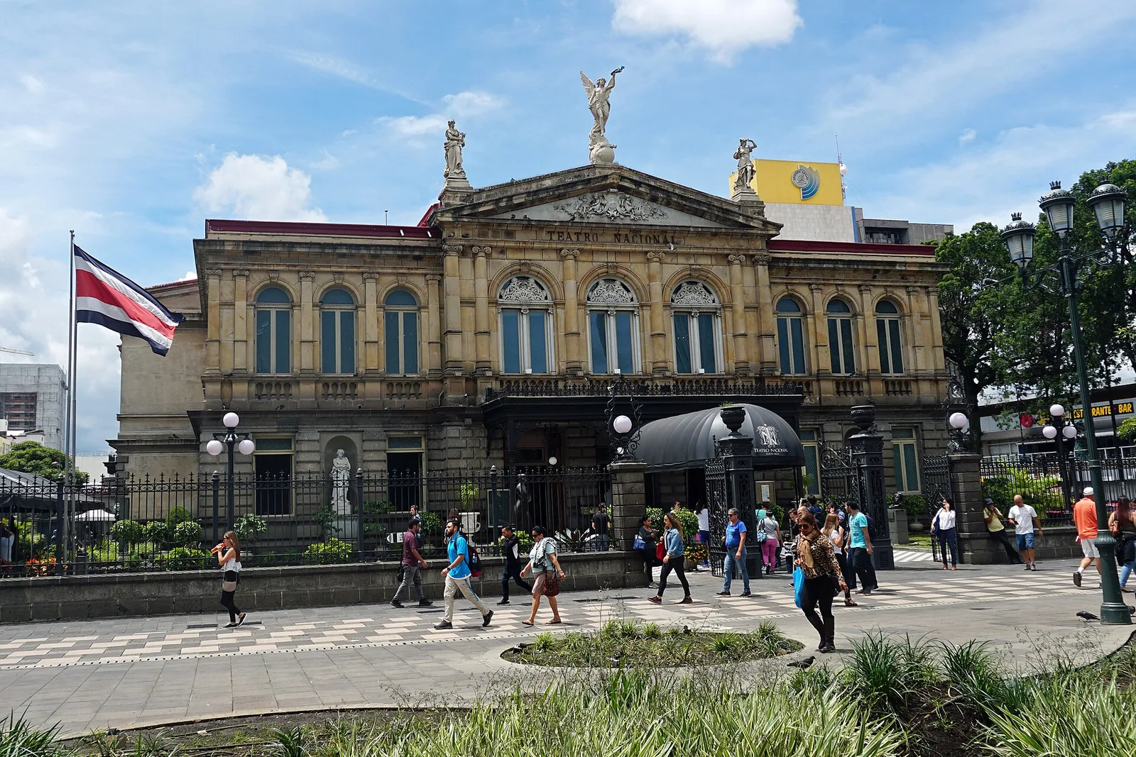 The neoclassical facade of the National Theater of Costa Rica across a paved plaza, with a flag at the left