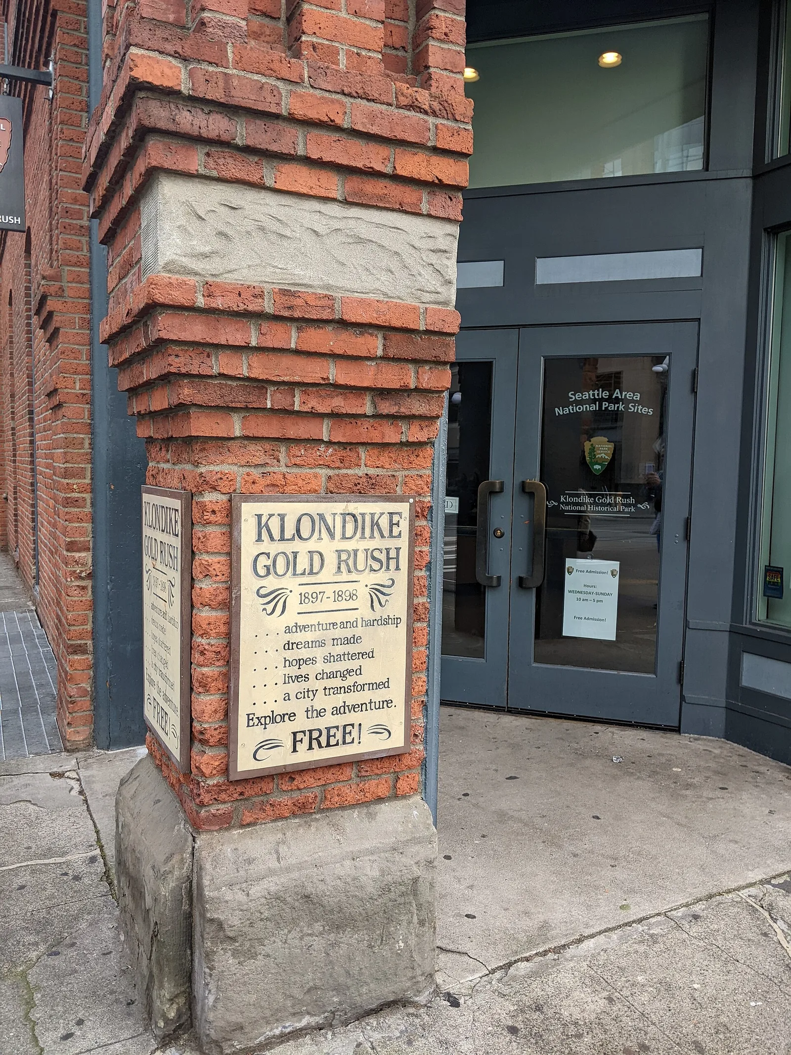 The brick corner of the Klondike Gold Rush museum building in Pioneer Square, with a plaque reading “Klondike Gold Rush 1897–1916” and the park service entrance beside it
