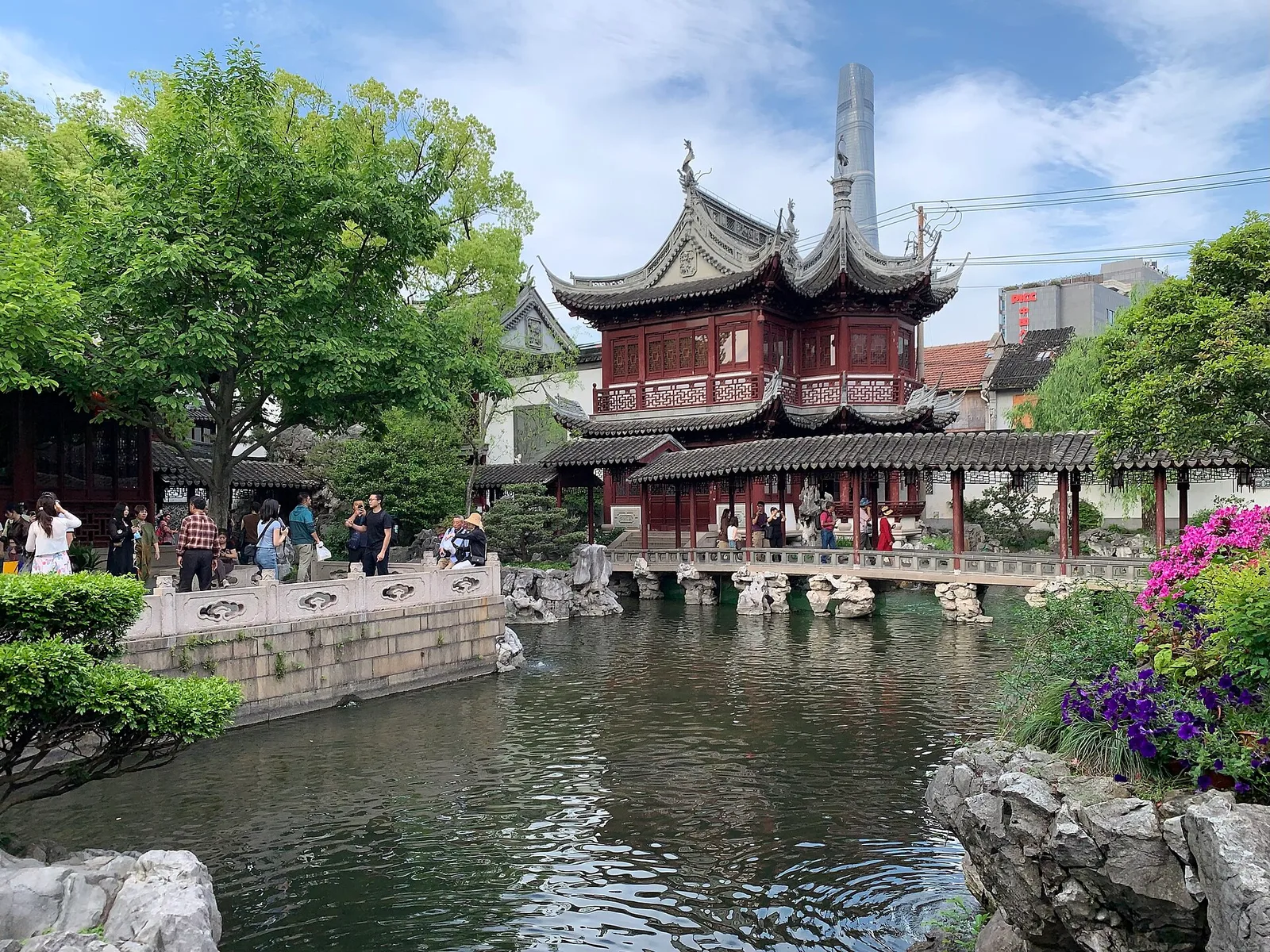 A red-columned pavilion with upturned eaves beside a pond in Yu Garden, with a skyscraper behind