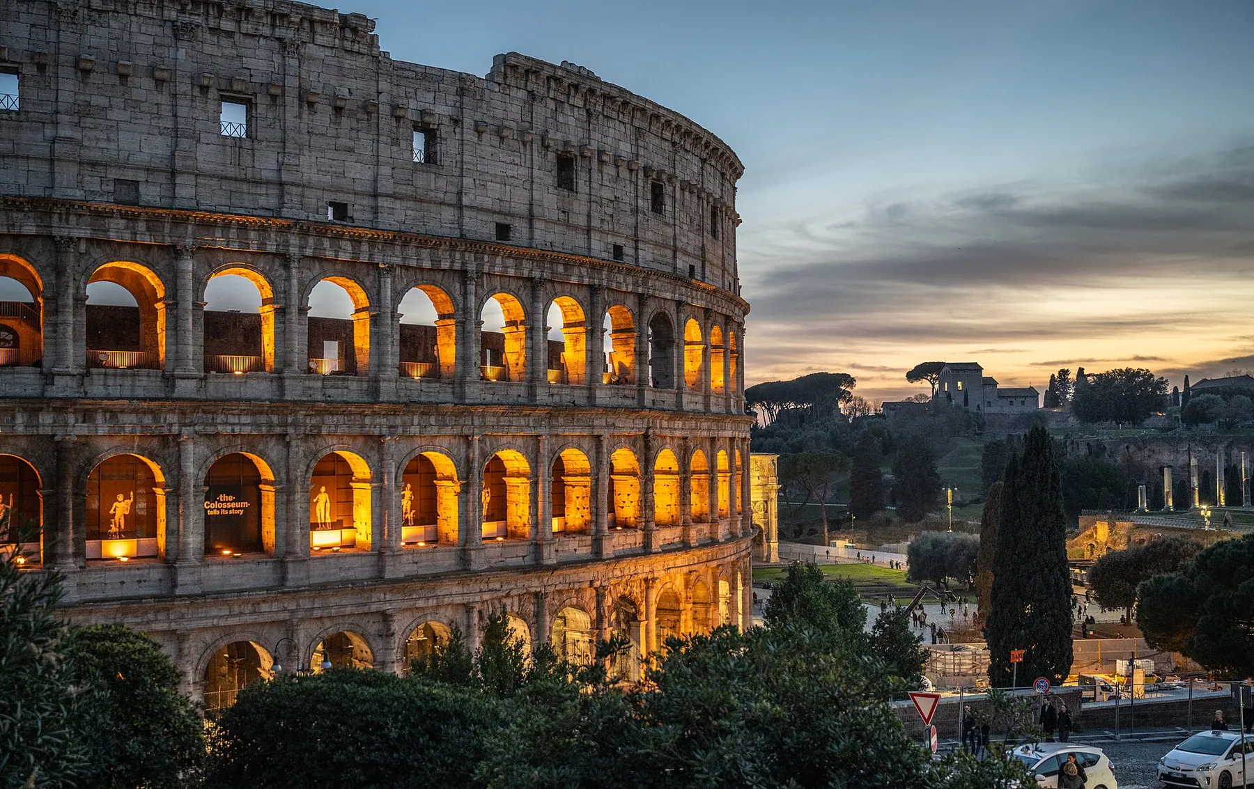 The Colosseum at dusk with its arches lit warm orange against a pale evening sky