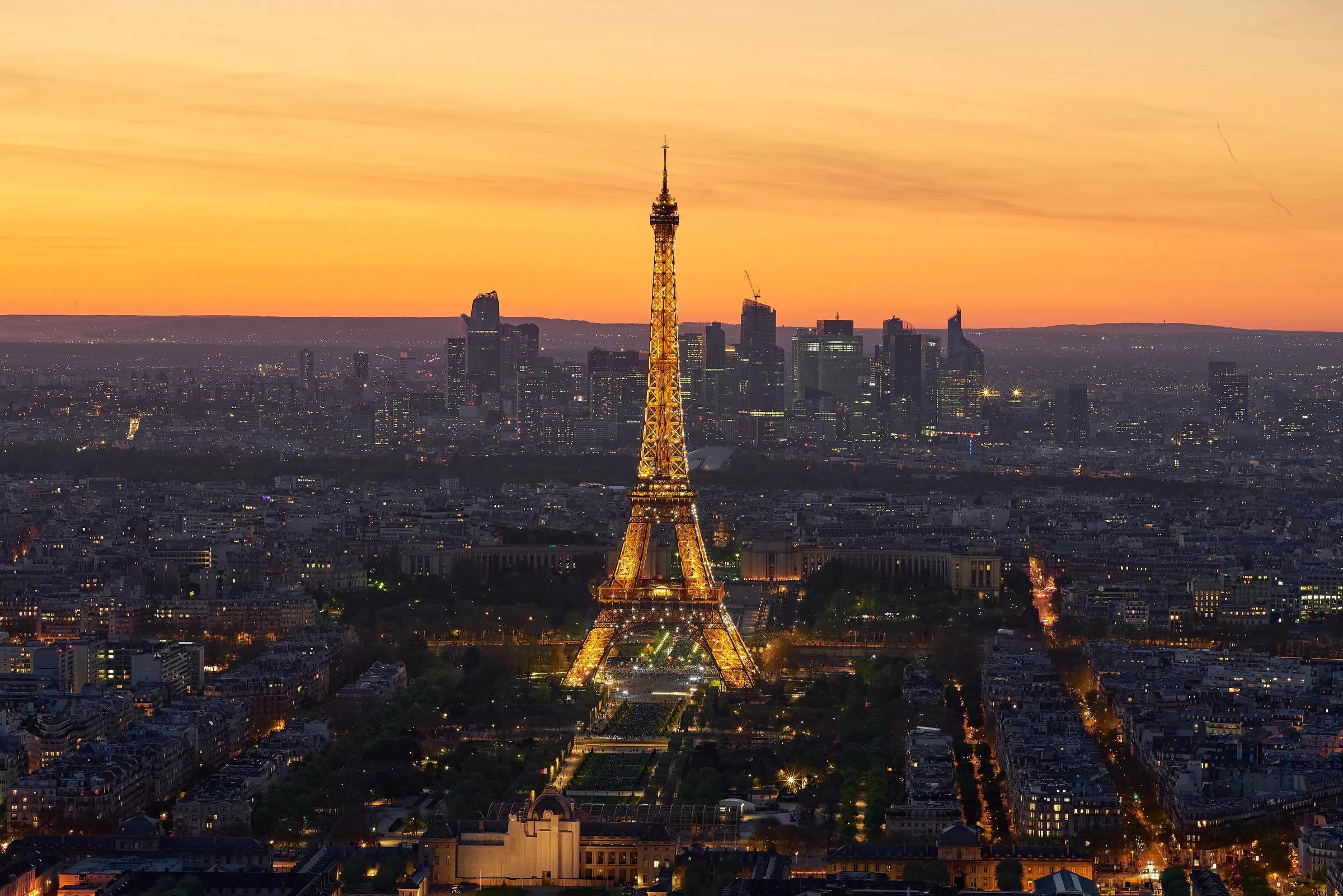 The Eiffel Tower lit up against an orange sunset sky above the rooftops and towers of Paris